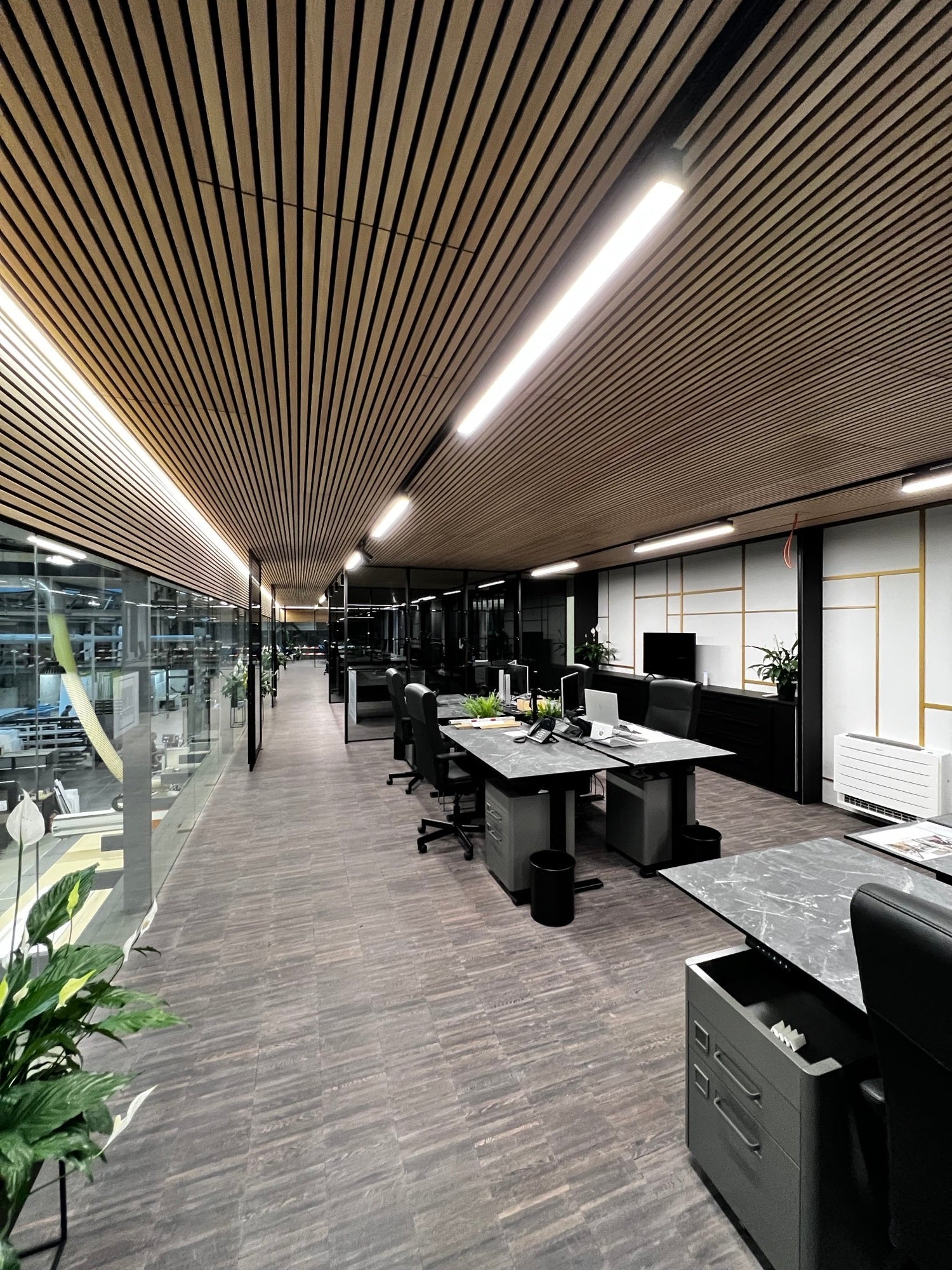 Modern kantoor met een Akupanel-plafond van houten latten, een glazen wand, minimalistische bureaus met computers, zwarte stoelen, planten en een op de Scandinavische stijl geïnspireerde inrichting van de werkruimte.